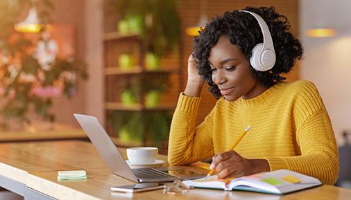 带着无线耳机微笑的黑人女孩在网上学习，在咖啡馆用笔记本电脑，做笔记